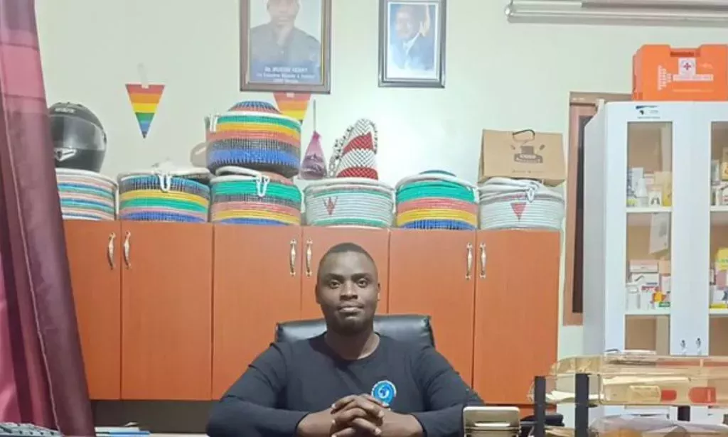 Henry Mukiibi, an LGBTQ+ activist from Uganda, wears a dark shirt as he sits at a desk with several items behind him