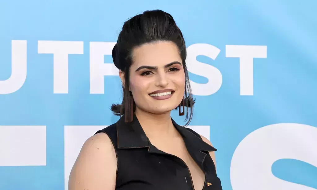 Nava Mau wears a black dress and smiles while standing against a blue background.