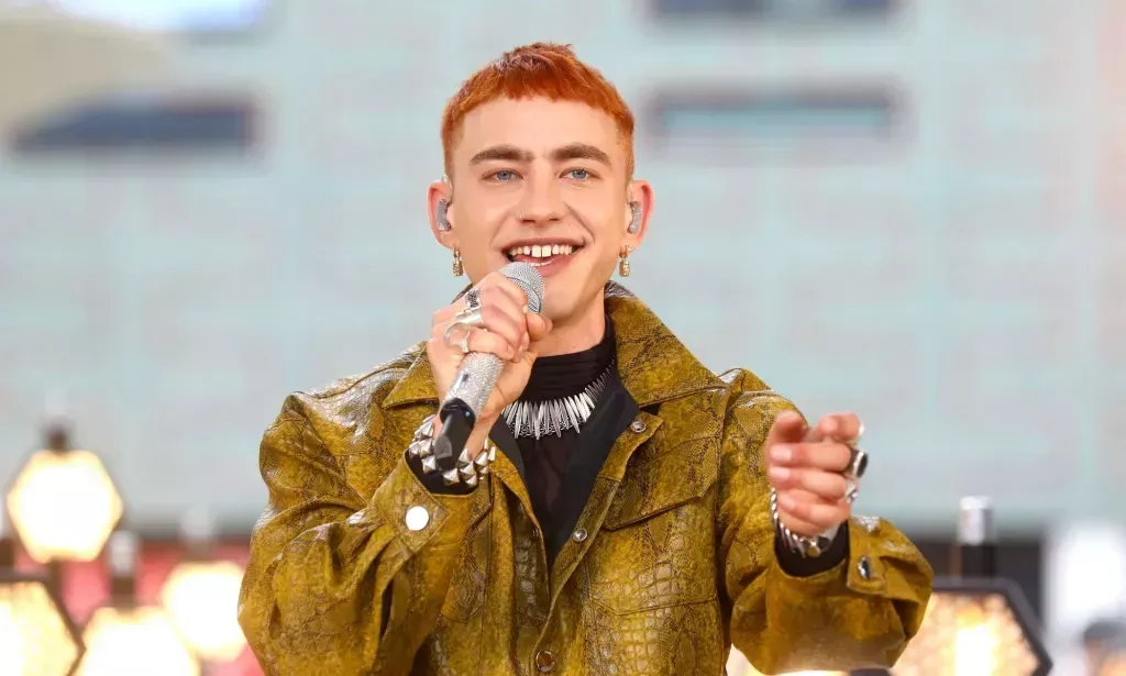 Olly Alexander in a yellow/green snakeskin jacket holding a microphone up to his mouth.