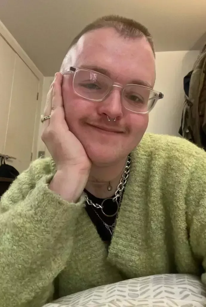 Kyle Cook, an activist living with HIV and drag queen, wears a dark shirt and green jumper as he smiles towards the camera