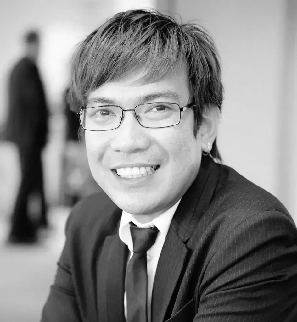 This is a black and white portrait of an Asian man. He is wearing wire rimmed glasses and has shaggy short hair. He is wearing a black suit.