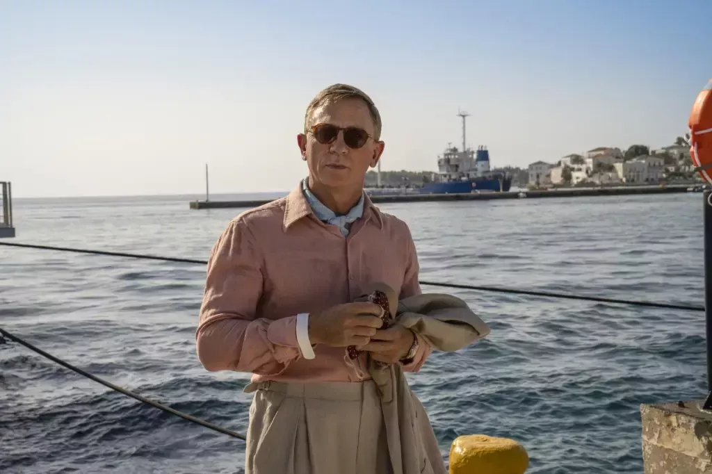 Daniel Craig in Glass Onion: A Knives Out Mystery, he's standing in a pink shirt and nude trousers by the side fo the ocean.