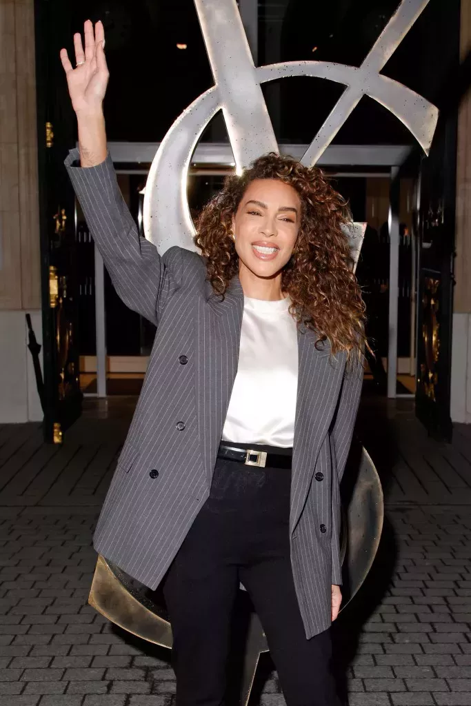 Ines Rau in front of a metal YSL statue smiling and waving. She's wearing a pinstripe grey blazer, white top and black trousers with curly styled hair.