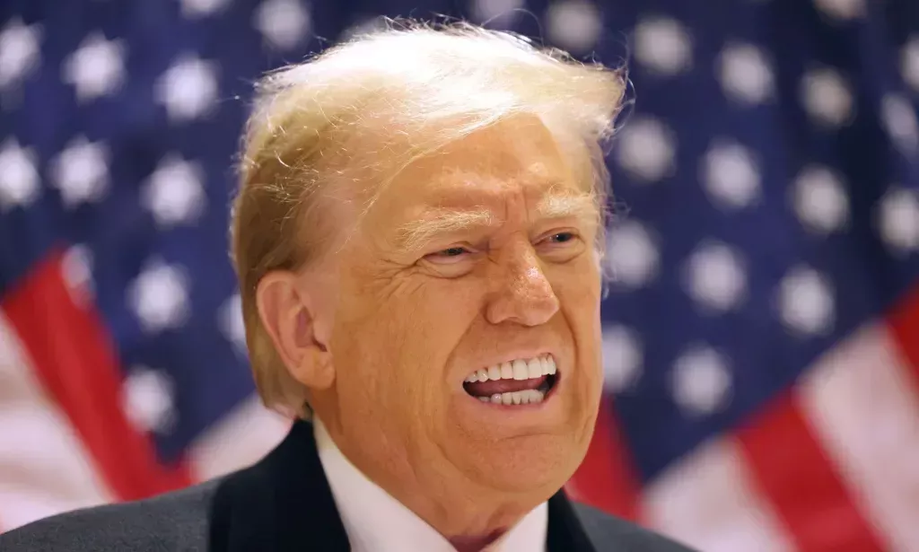 Former president Donald Trump wears a suit and tie as he speaks to people off camera and stands in front of red, white and blue US flags