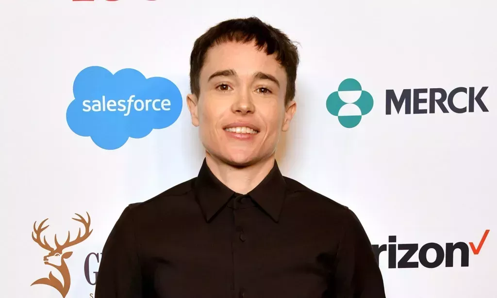 Elliot Page smiles on a red carpet while wearing a dark buttoned shirt.