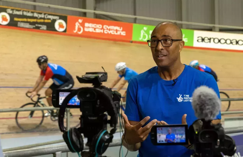 Colin Jackson at the Birmingham 2022 Commonwealth Games, commentating with the camera on him as three cyclists pass by behind him.