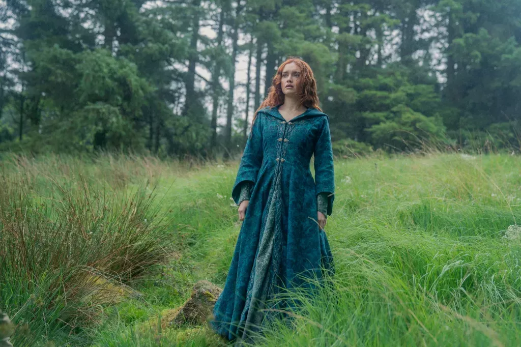 Alicent in a long dark green dress-coat walking through the woodland