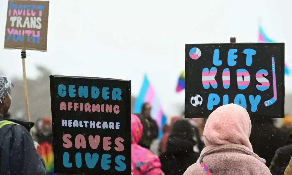 Trans people and allies hold up signs calling for protections for gender-affirming healthcare (including puberty blockers), trans youth and trans inclusion in sports during protest