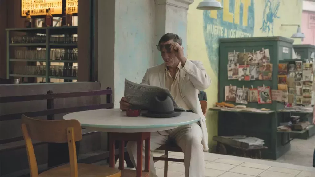 Queer still, a man is sat in a cream suit, removing his sunglasses as he reads the paper