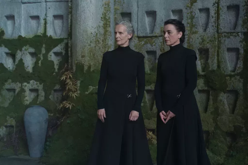 Barbra Marten y Olivia Williams en Dune: Profecía. Vestidas de negro completo contra una pared musgosa.