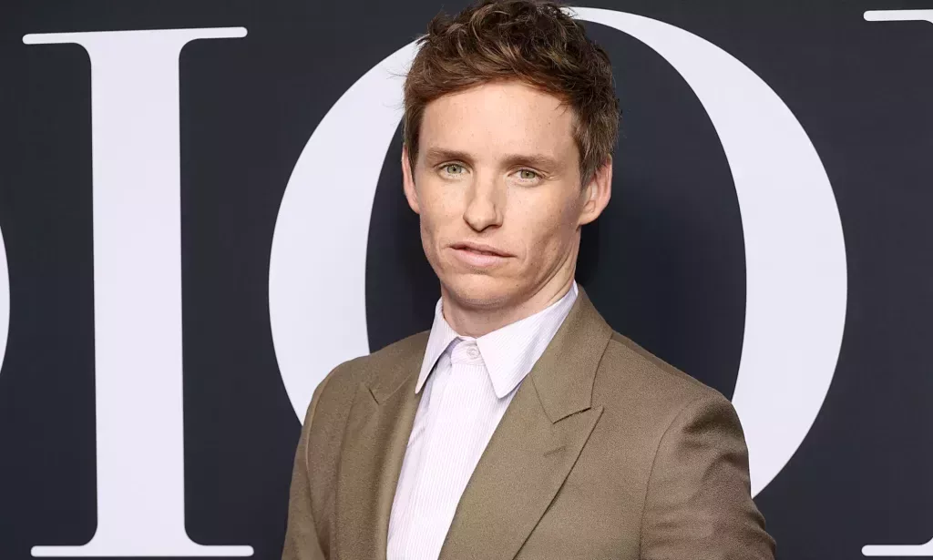 Eddie Redmayne lleva una camisa blanca con un abrigo marrón claro mientras posa para la cámara en un evento