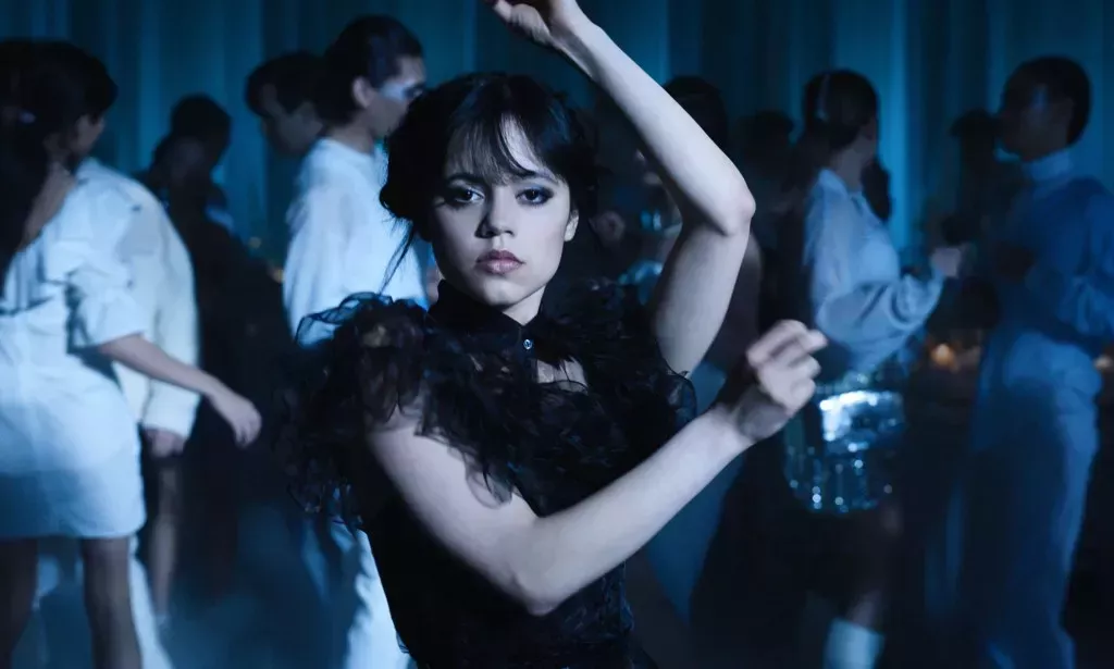 Una captura de pantalla de Wednesday de Netflix muestra a Jenna Ortega como Wednesday Addams vestida de negro sosteniendo una pose mientras baila - con otras personas también visibles en el fondo bailando.