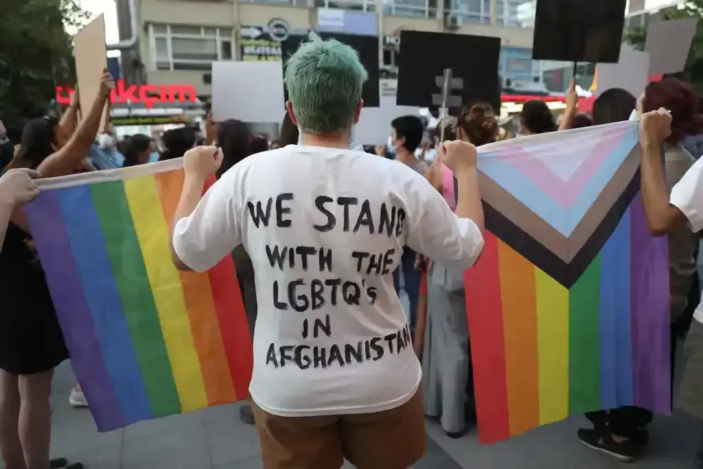 Persona de espaldas. La parte trasera de su camiseta dice: 'Apoyamos a los LGBTQ en Afganistán'. Está sosteniendo una bandera del Orgullo y la bandera del Orgullo Progresivo