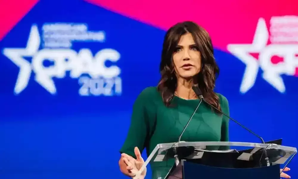 Kristi Noem usa un atuendo verde y está de pie en un podio frente a un fondo rojo y azul