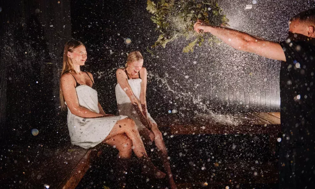 Un maestro de sauna agitando hojas a dos personas sentadas en una sauna estonia.