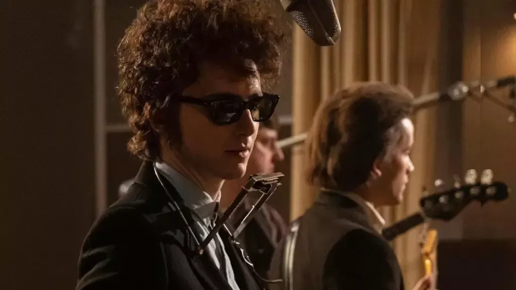 A Complete Unknown still: Timothée Chalamet como Bob Dylan en una grabación de estudio. Tiene un gran cabello rizado, gafas de sol oscuras y un atril para armónica alrededor de su cuello.