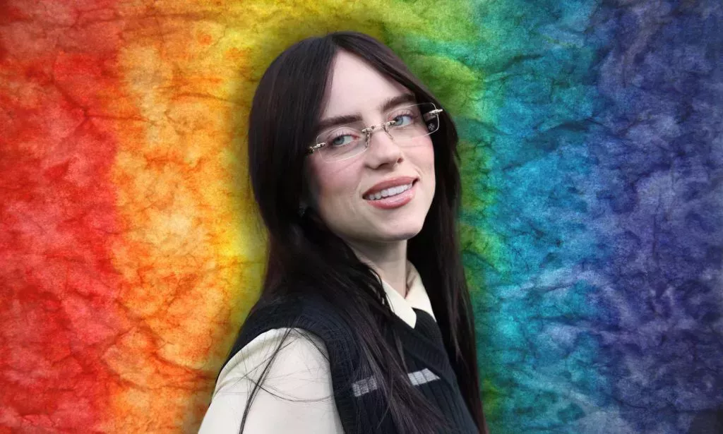 Billie Eilish mira hacia la derecha y sonríe. Ella está usando gafas y una camisa beige con un chaleco negro. Está contra un fondo arcoíris.